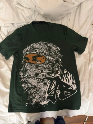 Grön t-shirt med grafiskt tryck - Mörkgrön t-shirt med stort grafiskt tryck framtill av en maskerad person och graffiti-inspirerad text med eldflammor. Baksidan har också ett stort tryck med samma text och eld. Klassisk passform och rund hals. Perfekt för dig som gillar streetwear och unika prints.