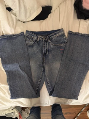 Blå bootcut jeans med slitningar - Säljer ett par blå bootcut jeans med lätt slitna detaljer och två röda märken vid fickan. Jeansen har klassisk femficksdesign, normalhög midja och är tillverkade i denim. Perfekta för en avslappnad och trendig look.