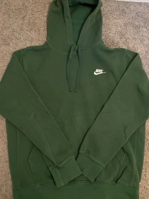Nike hoodie - Mörkgrön hoodie från Nike med klassisk vit broderad logga på bröstet. Tröjan har huva med dragsko och en stor magficka framtill. Tillverkad i mjukt material som känns skönt mot huden. Perfekt för en avslappnad och sportig stil.