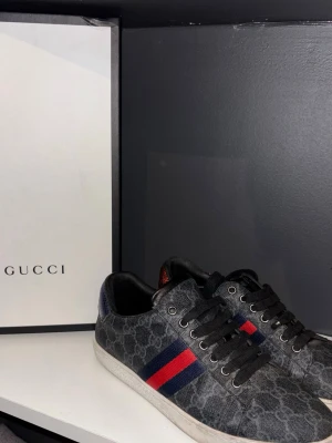Gucci sneakers med monogram och ränder - Snygga sneakers från Gucci i svartgrått med klassiskt GG-monogram över hela skon. Skorna har svarta snören och de ikoniska röda och blå ränderna på sidan. Låg modell med vit sula och metallfärgade öljetter. Perfekta för dig som vill sticka ut med lyxig streetstyle.