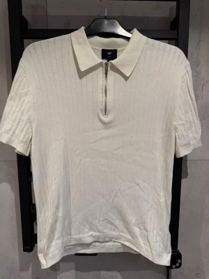 Vit ribbad polotopp med dragkedja - Säljer en stilren vit zip polotopp från H&M med korta ärmar och ribbad struktur. Toppen har en klassisk krage och en dragkedja framtill som ger en modern touch. Perfekt för dig som gillar enkel men snygg stil. 