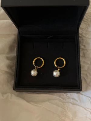 Guldiga örhängen med pärlor - Säljer ett par så såå söta stilrena örhängen med hängande vita pärlor och guldplätering. Örhängena har en klassisk rund form och pärlorna ger en elegant touch. Perfekta för dig som gillar tidlös och enkel design. Ej använda. Nypris 259!