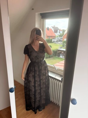Svart långklänning med spetsdetaljer - Svart långklänning med v-ringning och korta ärmar. Klänningen har ett transparent spetslager med blommigt mönster och markerad midja. Perfekt för dig som vill ha en elegant och romantisk look. Aldrig använd. OBS jag är en M så är ej rätt representant för klänningens passform för en L . Dock är den stretchig i midjan och har en dragkedja i ryggen. 
