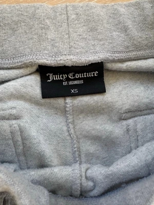 Gråa mjukisbyxor Juicy Couture XS - Mysiga gråa mjukisbyxor från Juicy Couture i storlek XS. Byxorna har raka ben, bred resår i midjan med dragsko och stora fickor framtill. Tillverkade i mjukt velourmaterial som känns lyxigt och skönt. Perfekta för chill dagar hemma eller när du vill vara extra bekväm.