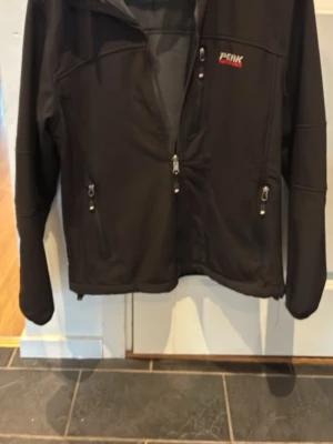 Svart vindjacka från Peak Performance - Snygg svart jacka med dragkedja framtill och två fickor med dragkedja. Jackan har en enkel, sportig design.  Perfekt för blåsiga dagar och outdoor-aktiviteter.
