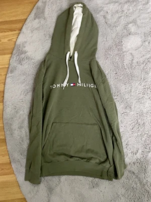 Olivgrön hoodie Tommy Hilfiger  - Snygg olivgrön hoodie från Tommy Hilfiger med vit logga och flagga på bröstet. Hoodien har stor huva med vita snören och en klassisk magficka framtill. Perfekt för en avslappnad och trendig look.