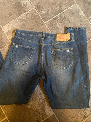 Levi's 501 blå jeans W36 L34 - Klassiska blå Levi's 501 jeans med rak passform och fem fickor. Jeansen har en snygg sliten look med en liten reva på bakfickan och tydlig Levi's-lapp bak i midjan. Tillverkade i kraftig denim med den ikoniska röda Levi's-taggen.