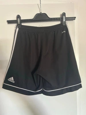 Svarta Adidas shorts med vita ränder - Svarta Adidas shorts med vita ränder längs sidorna och vit kant nertill. Klassisk sportig look med elastisk midja och diskret logga på ena benet. Perfekta för träning eller chill dagar. Tillverkade i polyester för en lätt och luftig känsla.