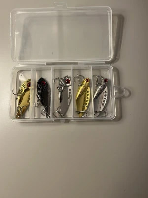 Fiskedrag i metall med ask - Säljer ett set med fem fiskedrag i metall, både i guld- och silverfärg. Dragena har röda detaljer vid ögonen och är utrustade med trekrokar. Kommer i en genomskinlig plastask med fack för varje drag. Perfekt för fiskeentusiasten som vill ha variation.