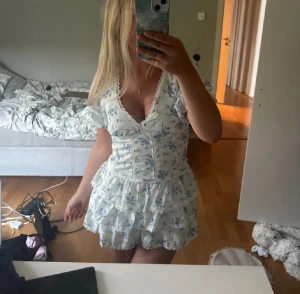Vit blommig kortklänning med volanger - Säljer denna fina klänning då den aldrig kommer till användning, helt oanvänd!