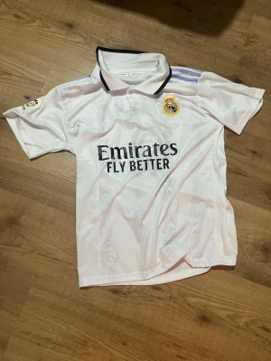 Real Madrid matchtröja Benzema 9 - Säljer en vit Real Madrid matchtröja med svart krage och lila detaljer. Tryck med 'Emirates Fly Better' på framsidan och 'Benzema 9' på ryggen. Klubbmärke på bröstet och La Liga-märke på ärmen. Perfekt för fotbollsfans!