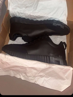 Svarta sneakers med randig sula - Snygga helsvarta sneakers med låg profil och unik randig detalj på sulan. Skorna har en slimmad design och är tillverkade i ett material som ser ut som syntetläder med meshdetaljer vid snörningen. Perfekta för dig som gillar stilrena och moderna skor.