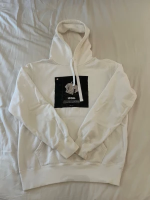 Vit hoodie med tryck och huva - Säljer en vit hoodie med stor huva och dragsnören. På bröstet finns ett svartvitt tryck med texten 'ICON.' och en grafisk bild. Hoodien har känguruficka framtill och ribbade muddar vid ärmar och nederkant. Perfekt för en chill och stilren look.