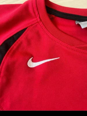 Röd Nike sporttröja med svarta detaljer - Snygg röd sporttröja från Nike med svarta paneler på sidorna och vit Nike-logga på bröstet. Tröjan har rund halsringning och korta ärmar, perfekt för träning eller match. Tillverkad i ett lätt och funktionellt material som andas.