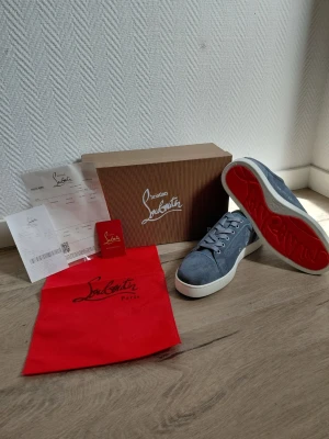 Christian Louboutin - Snygga gråa sneakers från Christian Louboutin i mjuk mocka med klassisk röd sula och vita kanter. Skorna har lågt skaft, matchande grå skosnören och diskreta sömmar för en clean look. Perfekta för dig som vill sticka ut med exklusiva detaljer.
