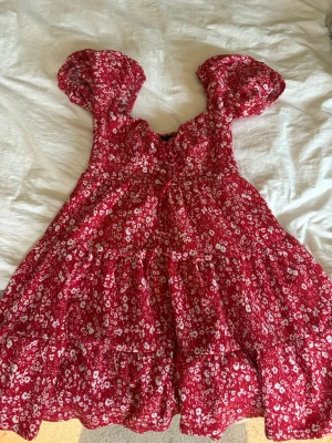 Röd blommig kortklänning med puffärm - Supersöt röd kortklänning med vita blommor och puffärmar. Klänningen har en luftig passform och volangdetaljer, perfekt för varma dagar. Den har en romantisk vibe och är tillverkad i ett lätt material som känns skönt mot huden.