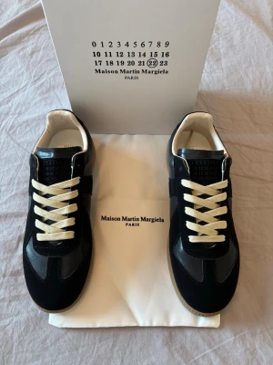 Maison Margiela svarta sneakers - Svarta sneakers från Maison Martin Margiela med vita snören och beige innersula. Skorna har en klassisk låg siluett, ovandel i mocka och läder samt gummisula. Perfekta för dig som gillar stilrena och tidlösa sneakers med exklusiv känsla.