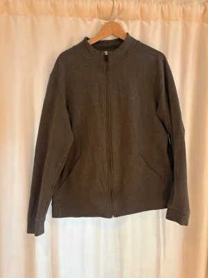 Mörkgrå zip hoodie från BOSS - Mörkgrå hoodie med dragkedja från BOSS. Tröjan har en enkel och stilren design med två fickor framtill och ribbade muddar vid ärmar och nederkant. Tillverkad i mjukt sweatshirtmaterial, perfekt för en avslappnad look. Sitter som en L