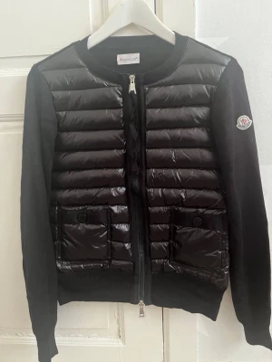 Svart cardiganjacka från Moncler - Snygg svart cardiganjacka från Moncler med quiltad framsida i glansigt material och stickade ärmar. Jackan har två framfickor med knappar, rund halsringning och dragkedja framtill. Moncler-logga på ena ärmen. Perfekt för lager på lager. Priset går att diskutera vid snabb affär, finn de frågor så är de bara o skriv 😊