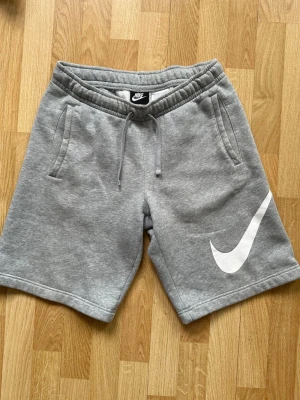 Gråa Nike Shorts - Säljer ett par gråa shorts från Nike. Shortsen har två sidofickor, en bakficka och en stor vit Nike-logga på ena benet. Perfekta för chill dagar eller träning.