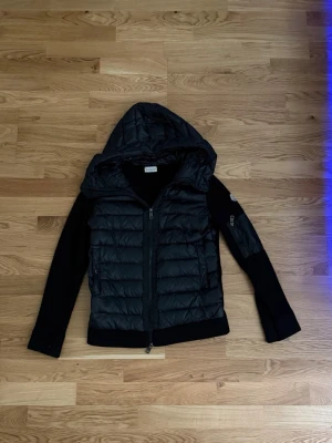 Svart jacka från Moncler - Snygg svart pufferjacka från Moncler med vadderad kropp och stickade ärmar. Jackan har en stor huva, dragkedja framtill och Moncler-logga på ärmen. Perfekt för kalla dagar och riktigt trendig look. M men passar också S