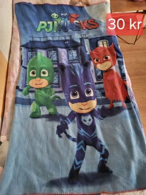 PJ Masks filt med motiv - Mjuk filt med färgglatt PJ Masks-motiv. Filtens framsida har tryck av karaktärerna Gecko, Catboy och Owlette i grönt, blått och rött mot en blå bakgrund. Perfekt för soffmys eller som barnrumsdekoration. Tillverkad i fleece-liknande material.