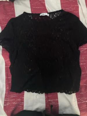 Svart spetsig kortärmad topp - Säljer en svart kortärmad topp med spetsdetaljer och lätt transparent material. Toppen har en rund halsringning och vågig kant nertill samt vid ärmslut. Perfekt för dig som vill ha en edgy och trendig look.
