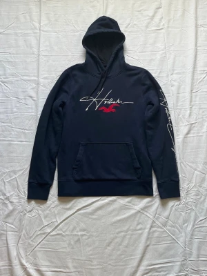 Mörkblå hoodie från Hollister - Snygg mörkblå hoodie från Hollister Must-Have Collection med vit broderad logga och röd fågel på bröstet. Stor ficka framtill, huva med dragsko och tryck även på ena ärmen. Perfekt för en avslappnad och trendig stil.