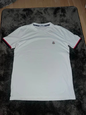 Vit Moncler t-shirt med logga - Säljer en stilren vit t-shirt från Moncler med rund hals och korta ärmar. T-shirten har Moncler-logga broderad på bröstet och snygga detaljer i rött och blått på ärmsluten. Perfekt för dig som gillar klassisk och enkel design.
