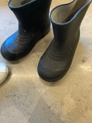 Svarta gummistövlar med foder - Svarta höga gummistövlar med grått innerfoder. Klassisk design med rund tå och räfflade detaljer framtill. Perfekta för regniga dagar och leriga äventyr. Tillverkade i slitstarkt gummi med mjukt textilfoder för extra komfort.