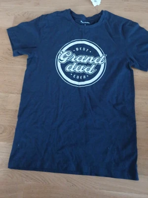 Mörkblå t-shirt Granddad Kappahl - Mörkblå t-shirt från Kappahl med trycket 'Best Granddad Ever' i vitt på bröstet. Klassisk rund hals och korta ärmar. Tillverkad i mjuk bomull för skön känsla och enkel stil.