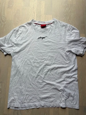 Vit t-shirt från Hugo med broderad logga - Säljer en vit t-shirt från Hugo med svart broderad logga på bröstet. Klassisk rund halsringning och korta ärmar. Tillverkad i mjuk bomull som känns skön mot huden. Perfekt för en clean och stilren look. Ny pris runt 450kr