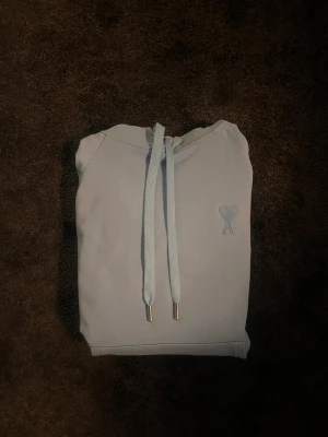 Ljusblå Ami Paris hoodie  - Säljer en ljusblå hoodie med huva och dragsnören med metalländar. På bröstet finns en liten broderad hjärtdetalj. Hoodien har en klassisk känguruficka framtill och ribbade muddar vid ärmar och nederkant. Perfekt för en avslappnad och stilren look.