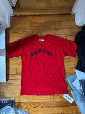 Röd t-shirt med svart tryck - Snygg röd t-shirt med svart texten 'spöder' på bröstet. T-shirten har små glittriga prickar över hela tyget och klassisk rund hals. Perfekt för dig som vill sticka ut med en unik design.