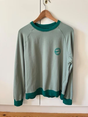 Grön crewneck med tryck från Bonprix strl 48/50 M - Ljusgrön sweatshirt från Bonprix bpc collection med mörkgröna muddar och krage. Tröjan har ett litet tryck på bröstet med text och symbol. Material 95% bomull, 5% elastan. Storlek 48/50, M.