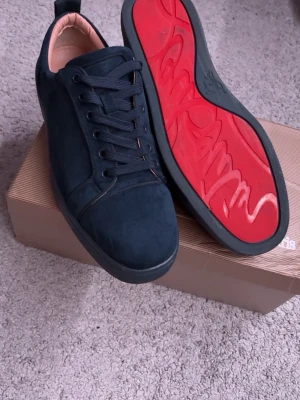 Loubs skor med röd sula - Använda 5 gånger.Snygga svarta sneakers i mocka med klassisk låg profil och svarta skosnören. Insidan är ljusbeige och yttersulan är svart med en cool röd undersida och grafiskt mönster. Perfekta för dig som vill ha en stilren men unik look.