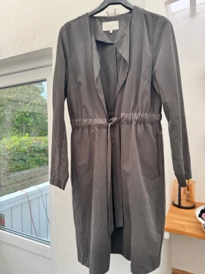 Brun trenchcoat/coatigan från Vila - Stilren brun trenchcoat/coatigan från Vila med knytband i midjan och långa ärmar. Jackan har öppen front utan knappar och ett mjukt, tunt tyg som ger ett avslappnat intryck. 