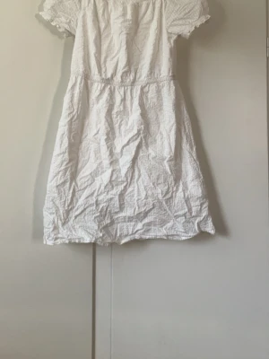 Vit kortärmad klänning med volang - Söt vit klänning med korta ärmar och volangdetaljer vid ärmslut och hals. Klänningen har en lätt rynkad struktur och är tillverkad i ett tunt, luftigt material som passar perfekt till varma dagar.