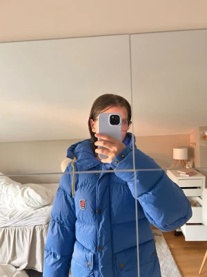 Blå dunjacka från Fjällräven - Snygg blå dunjacka från Fjällräven med gula dragskor och klassisk logga på bröstet. Jackan har knappar framtill, två fickor och en hög krage. Perfekt för kalla dagar och riktigt varm tack vare dunfyllningen. Jackan var min mammas förut och är i storlek S.