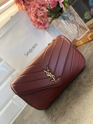 Vinröd YSL handväska i läder - Snygg vinröd handväska från Saint Laurent i quiltat läder med guldfärgad YSL-logga framtill. Väskan har kedjeremmar i guld och klassisk kuvertform. Perfekt för dig som vill ha en lyxig accessoar med ikonisk design.