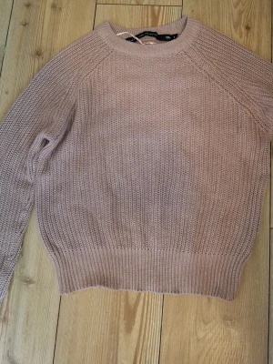 Ljusrosa stickad tröja - En mysig ljusrosa stickad tröja med rund halsringning och ribbade muddar vid ärmslut och nederkant. Tröjan har en relaxed passform och är perfekt för lager på lager under kyligare dagar.
