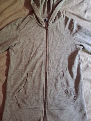 Ljusgrå zip hoodie med huva - En enkel och stilren ljusgrå hoodie med dragkedja framtill och två fickor. Hoodien har en klassisk huva och ribbade muddar vid ärmslut och nederkant. Perfekt för en avslappnad look och passar till det mesta. Materialet känns mjukt och bekvämt.