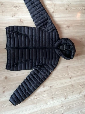 Svart dunjacka med huva från Colmar - Snygg svart dunjacka från Colmar med huva och två dragkedjeförsedda fickor framtill. Jackan har quiltad design och Colmar-logga på ärmen. Perfekt för kalla vinterdagar och riktigt lätt att bära. Materialet är glansigt och ger en modern look.