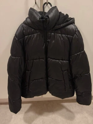 Svart glansig pufferjacka med huva - Snygg glansig svart pufferjacka med stor huva och dragkedja framtill. Jackan har en boxig passform, breda kanaler och två sidofickor. Jackan är från H&M och är i strl L. 