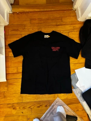 Svart t-shirt från Gallery Dept - Säljer en svart t-shirt från Gallery Dept med röd logga tryckt på bröstet. Klassisk rund hals och korta ärmar. Perfekt basic-plagg med streetwear-vibe som funkar till det mesta.