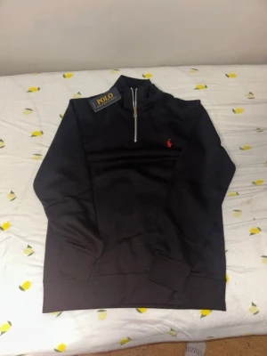 Svart half-zip tröja  - Svart half-zip sweatshirt med klassisk röd logga broderad på bröstet. Tröjan har hög krage, dragkedja och ribbade muddar vid ärmslut och nederkant. Perfekt för en clean och stilren look.