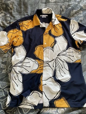 Blommig skjorta från Zara, slim fit - Säljer en snygg kortärmad skjorta från Zara i slim fit med stort blommigt mönster i vitt och gult på mörkblå botten. Perfekt för dig som vill sticka ut med en färgglad och trendig look. Skjortan har klassisk krage och knäppning framtill.