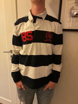 Randig rugbytröja med krage - Säljer en klassisk långärmad rugbytröja med breda marinblå och vita ränder. Tröjan har krage, knappslå och en röd 85-detalj på bröstet tillsammans med ett broderat emblem. Perfekt för en sportig och avslappnad stil.
