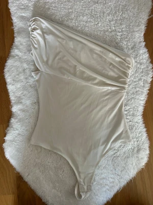 Vit oneshoulder body från H&M - Säljer en stilren vit body från H&M i oneshoulder-modell. Bodyn har en slät och mjuk känsla, tillverkad i stretchigt material som sitter snyggt på kroppen. Perfekt att matcha med jeans eller kjol för en trendig look.