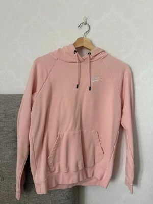 Ljusrosa hoodie från Nike - Säljer en ljusrosa hoodie från Nike med klassisk känguruficka och justerbar huva med snören. Tröjan har en liten vit Nike-logga på bröstet och är gjord i mjukt bomullsmaterial. Perfekt för en chill och avslappnad stil.
